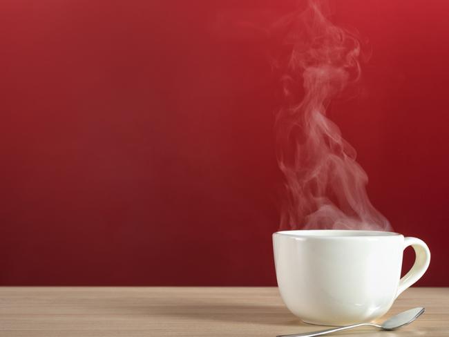 A hot steamy large white cup of coffee or tea with spoon on a blonde colored wood table against a red wall