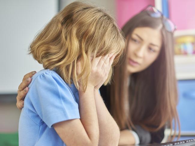 a primary school girl is sitting at her computer desk in school and crying into her hands . Her young teacher looks on  in the background , puts her arm around her and gives support .