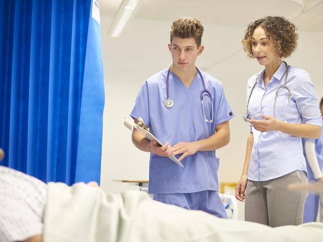 A young female doctor is chatting to a patient on a busy hospital ward with a male nurse  . The patient is unrecognisable , but in the background a  female nurse can be seen attending to other patients . The female doctor is wearing a shirt with rolled up sleeves and the male nurse is wearing scrubs. The hospital ward is similar to those used in a uk communal nhs hospital layout .