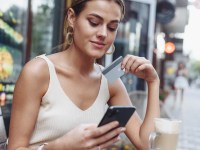 Woman sitting at cafe holding mobile phone and plastic card signing up on website.Beautiful model paying with credit card while shopping online using mobile.