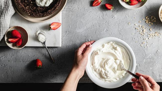 Pov shot of a female chef mixing yogurt in a bowl in kitchen with ingredients on kitchen counter. Woman preparing sweet pie using yogurt and strawberries.