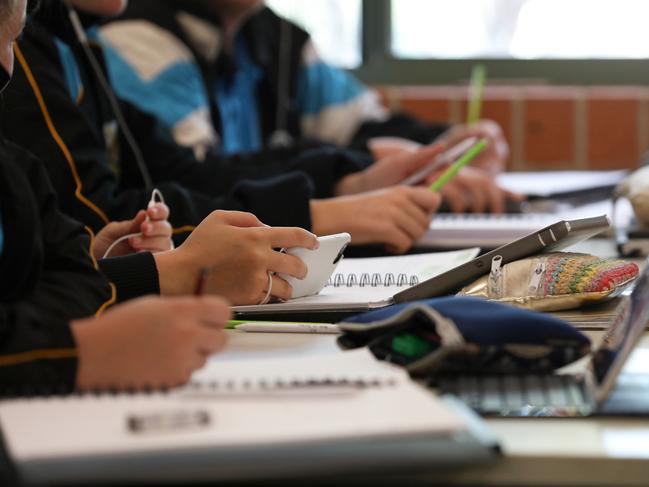 A student in uniform distracted holding using and watching a mobile phone during a lesson at high school. Books, tablets and pencil cases all visible on the desk and work space.