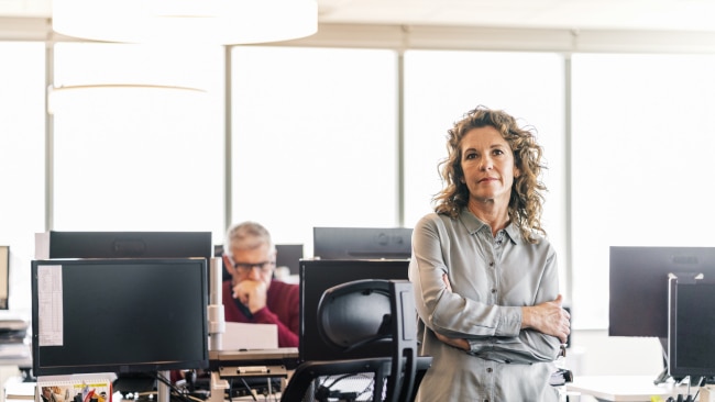 Thoughtful businesswoman standing with arms crossed. Mature professional is working on computers at desk. They are in office.