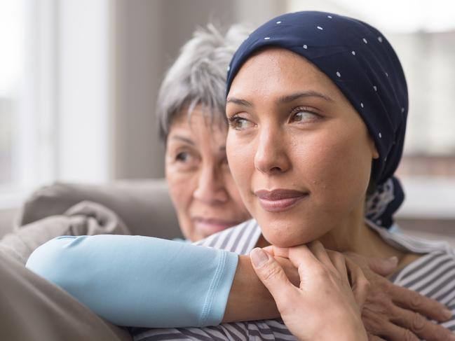 An ethnic woman wearing a headscarf and fighting cancer sits on the couch with her mother. She is in the foreground and her mom is behind her, with her arm wrapped around in an embrace, and they're both looking out the window in a quiet moment of contemplation.