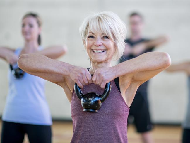 An older Caucasian woman is seen doing a squat with a kettlebell, while participating in a co-ed, multi-ethnic, fitness class. She is expressionless and focusing on her form. Active seniors generic