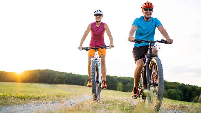 low angle view athletic sporty fit happy smiling woman and man couple friends in their fourties cycling with their electric mountain bicycles on gravel path through grassland rural landscape on sunny summer afternoon sunset