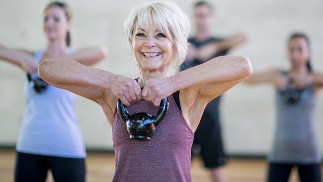 An older Caucasian woman is seen doing a squat with a kettlebell, while participating in a co-ed, multi-ethnic, fitness class. She is expressionless and focusing on her form. Active seniors generic