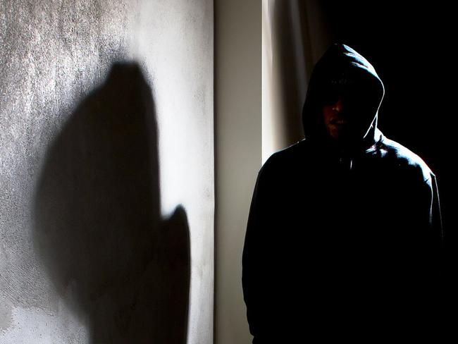 Photo of a hooded criminal stalking in the shadows of a dark street alley.  The hooded man is a silhouette and hiding in the dark.  The man is a criminal waiting to ambush victims.  The concrete walls provide copyspace.The photo depicts crime.