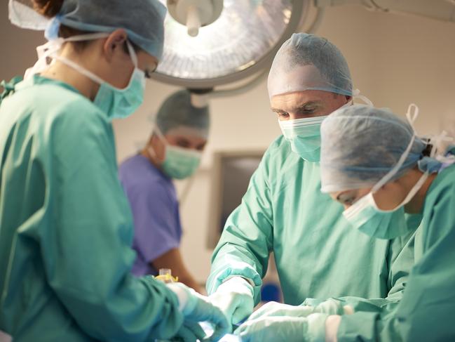 a female operating nurse stands over a patient at the operating table and looks down to what she is doing in the operation. She is joined by a young female nurse and mature male surgeon .In the background the anaesthetist is looking over from his monitors . They are all wearing surgical gowns and protective masks and headwear. they are wearing green gowns . The shot is horizontal waist up , with defocussed background for copy space .