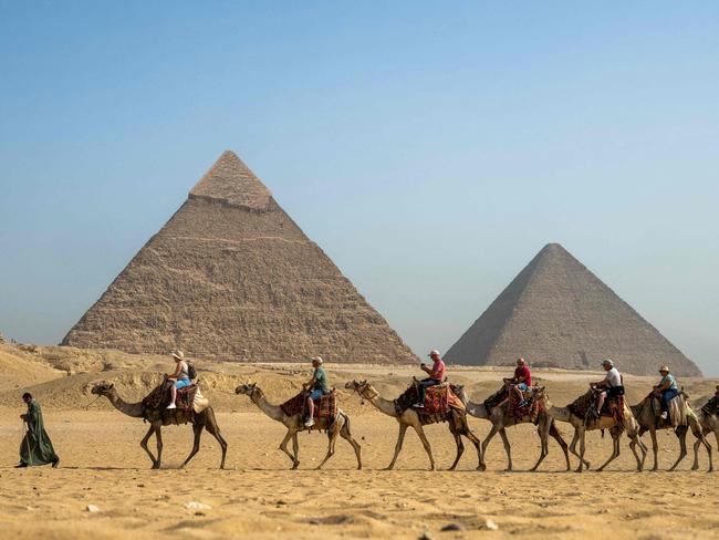 TOPSHOT - A guide pulls a lead camel as tourists ride in a row past the Great Pyramid of Khufu (Cheops, R) and the Pyramid of Khafre (Chephren, L) at the Giza Pyramids Necropolis on the outskirts of Giza on May 3, 2024. (Photo by Jewel SAMAD / AFP)