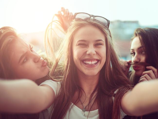 Group of teen friends how smiling, making faces and taking selfie with their mobile smartphone outdoor on the street at sunset.