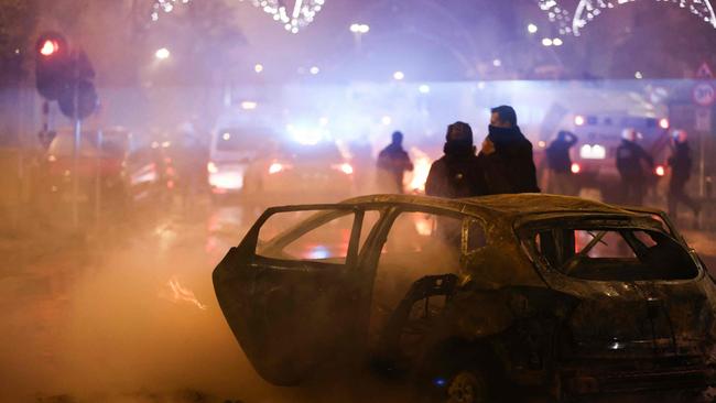 People stand next to a burnt car on the sideline of the live broadcast of the Qatar 2022 World Cup