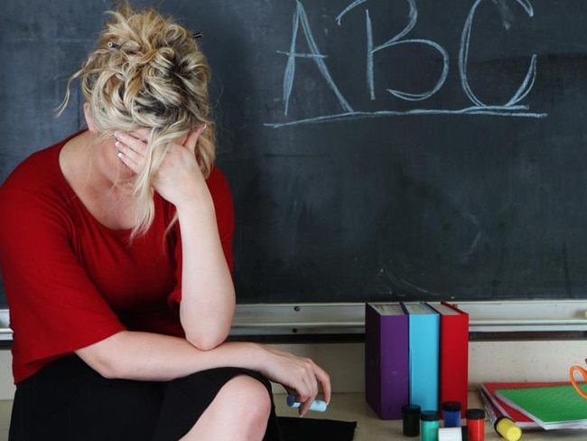 Elementary classroom setting with tired or frustrated teacher holding her head. She's sitting in front of an chalkboard with ABC