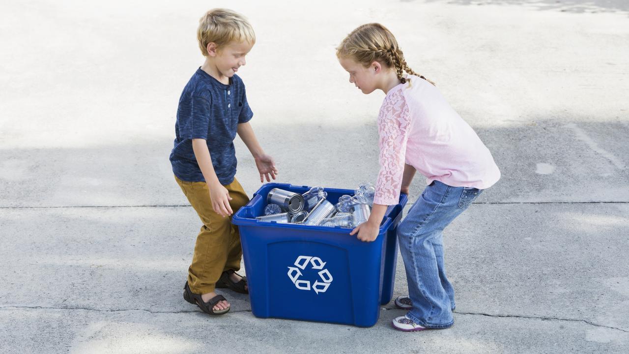 A young girl and her little brother carry a blue recycling bin full of empty bottles and cans to their curb for collection.