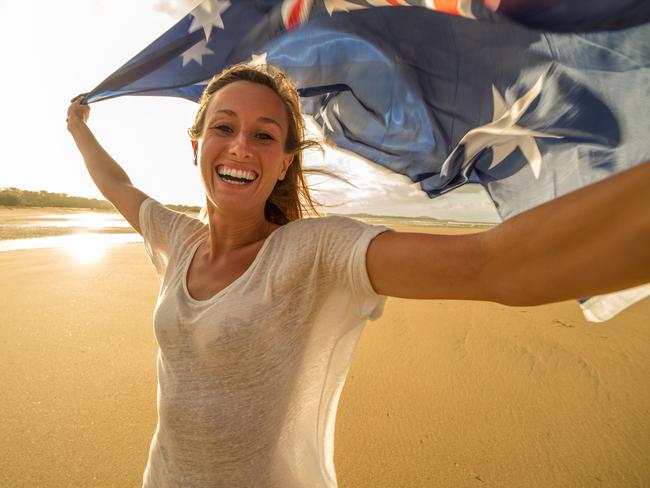 Young woman on the beach at sunset takes a selfie portrait while holding an Australian flag in the air.