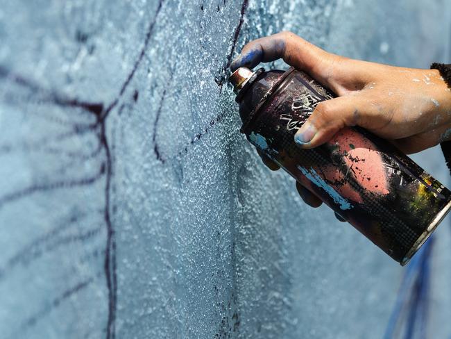 A young man holds a dark can of spray paint close to a blue wall to sketch his graffiti. iSTOCK