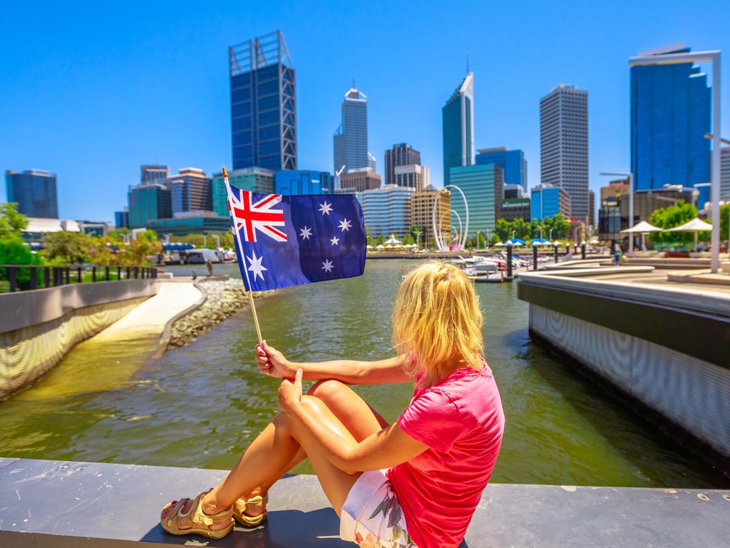Blonde woman with Australian flag on Elizabeth Quay Marina promenade in a sunny day. Central Business District in Perth, Western Australia on blurred background. Caucasian tourist enjoys city views.