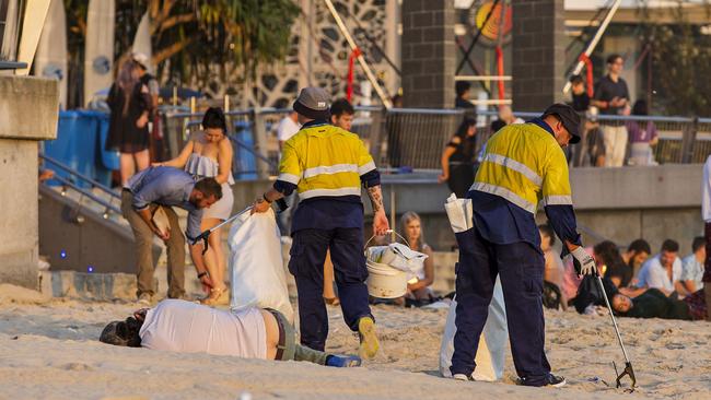 Two council workers cleaning rubbish of the beach as a man lays asleep next to them on the first day of 2017 at Surfers Paradise. Picture: Jerad Williams