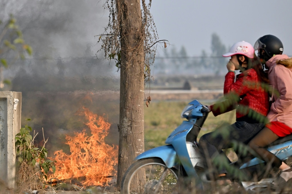 A woman covers her face as she rides a motorbike past a burning garbage dump on the side of the road in Hanoi