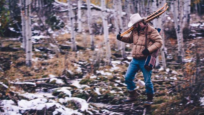 Man wearing cowboy hat carrying snowshoes over shoulder walks through snowy woods and forest while hiking and camping