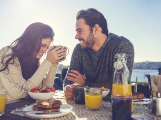 Couple eating breakfast outdoors. They are sitting on a patio or balcony eating muesli and toast and drinking coffee and juice. Sea in the background view