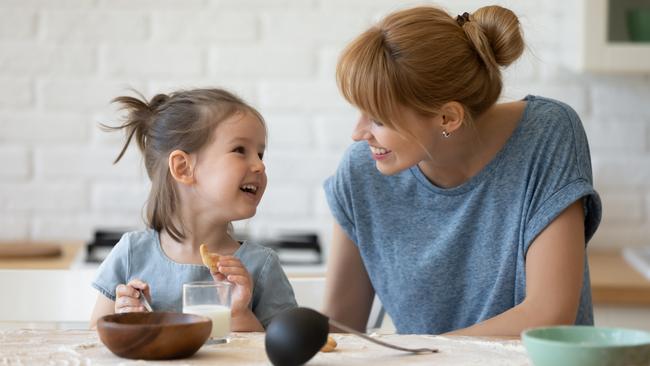 Smiling mother looking at little daughter and feeding her after bake cookies standing at table in kitchen. Happy caring mum and adorable girl eating flour and drink milk, preparing dinner.