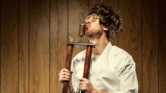 A nerdy looking guy wearing a white gi and black belt with big glasses and a goofy smile holds his new trophy he won at the competition, showing that he has mastered a martial art.   He stands in front of a wood paneled wall kissing the trophy with pursed lips; horizontal with copy space.