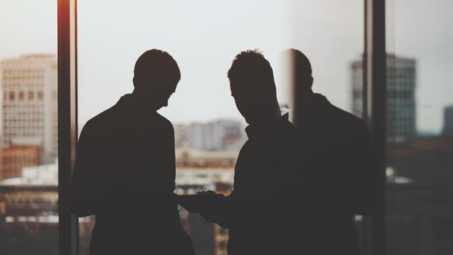 Silhouettes of two businessman near window and table of dark office interior, discussing something and looking on screen of digital tablet, cityscape outside