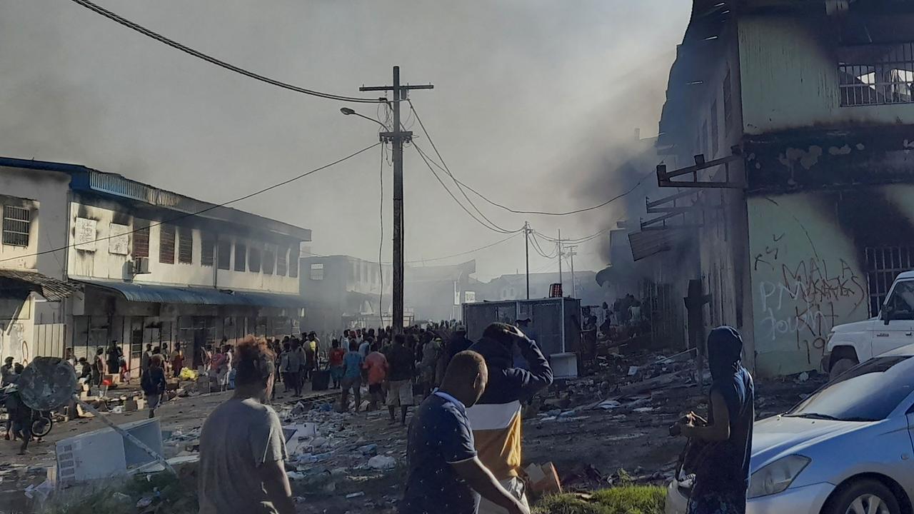 People gather as smoke rises from a burnt out buildings in Honiara's Chinatown on November 26, 2021 after two days of rioting. Picture: CHARLEY PIRINGI / AFP.