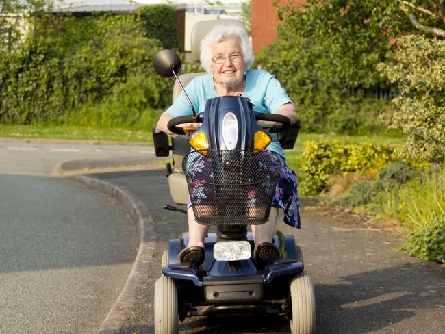 Elderly lady smiling happily as she rides her mobility scooter along the pavement.