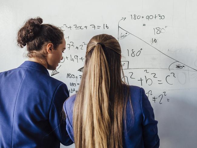 Two teenage school girls standing in front of  a large whiteboard side by side solving a mathematics equation on the board.