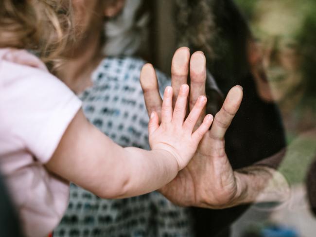 A mother stands with her daughter, visiting senior parents but observing social distancing with a glass door between them. The granddaughter puts her hand up to the glass, the grandfather and grandmother doing the same. A small connection in a time of separation during the Covid-19 pandemic.