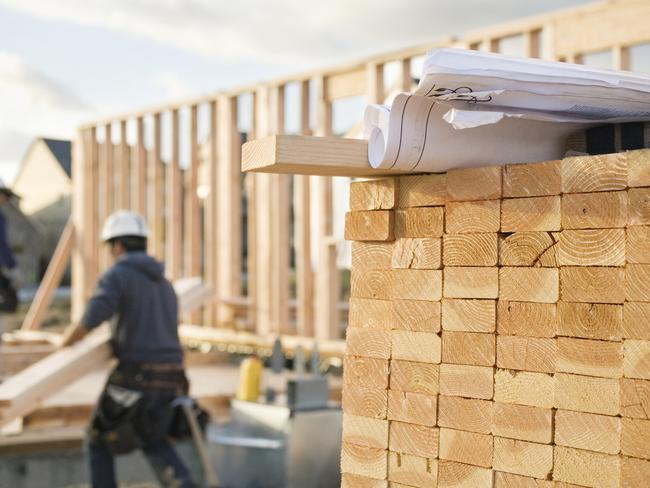 A closeup of stacks of 2x4 boards at a construction site, with a roll of blueprints sitting on top.  Two construction workers and a building frame can be seen in the background. Horizontal shot.