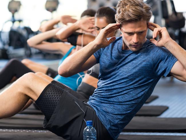 Muscular guy doing sit ups at gym with other people in background. Young athlete doing stomach workout in modern gym. Handsome fit man doing crunches at gym.