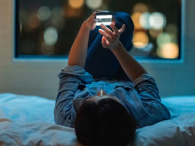 A woman is lying down on a bed and using a smart phone at night.Escape 22 October 2023Kendall HillPhoto - iStock