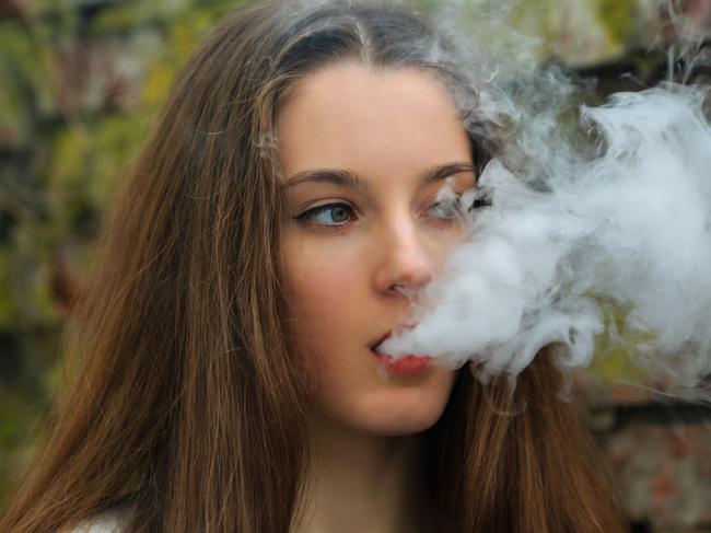 Vape teenager. Young pretty white girl in white cardigan smoking an electronic cigarette opposite destroyed brick wall on the street in the spring. Bad habit that is harmful to health.