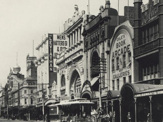 ooking south east along Bourke Street with Coles Book Arcade on opposite side of street, pavement in front of stores covered by porches with corrugated iron roofs, tram and horse drawn vehicles in street, Parliament House visible in distance. Please credit State Library of Victoria.