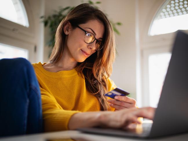 A beautiful young woman enjoying working at home on her laptop in cozy and bright apartment wearing yellow sweater and shopping online paying with credit card