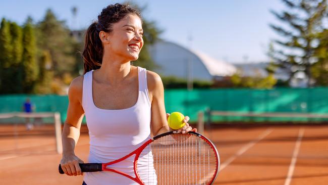 A woman with a radiant smile, holding a tennis racket on a clay court, posed gracefully against a vibrant backdrop of trees and clear skies during a sunny day.