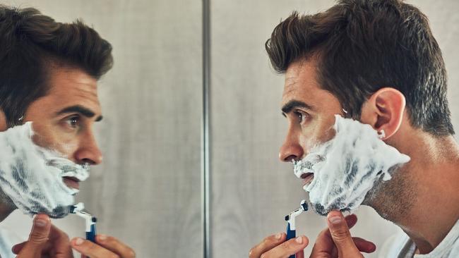 Shot of a focused young man shaving his beard while looking at his reflection in the mirror at home