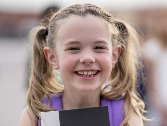 A sweet little girl with pigtails giggles outside as she poses for a portrait in front of her school.  She is dressed casually and is holding her books tightly as her peers play away in the background.