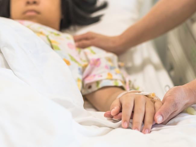 Nursing caretaker concept with kid patient sleeping in bed with family caregiver hand support in blur medical hospital background (focus on hand). Picture: iStock