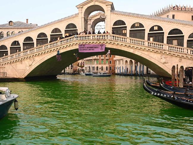 TOPSHOT - Extinction Rebellion (XR) activists are hanged on the Rialto bridge in Venice after fluorescein was poured into the Grand Canal to make the water fluorescent during a protest against the COP28 'fail' on climate, on December 9, 2023. The banner reads "COP28 : While the Government speak we are hanging on by a thread" (Photo by Marco SABADIN / AFP)