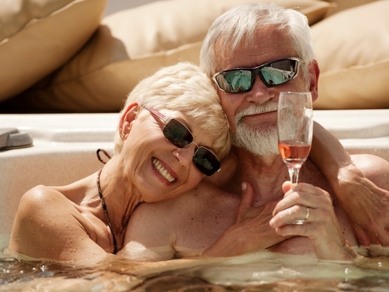 RendezView. Senior couple embracing and drinking sparkling wine in jacuzzi on the summer terrace. She has blond short hair and he has gray hair and beard. Both wearing sunglasses and smiling to each other. (Pic: iStock)