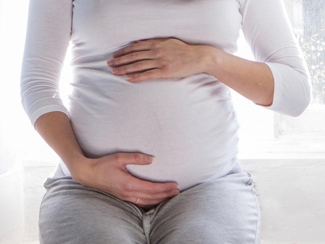 A heavily preganant woman is sat in her home bedroom. She is dressed in comfortable clothes, holding her baby bump.