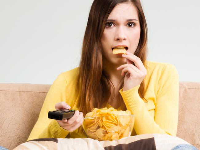 Young woman spends his free time watching TV on the couch at home, munching chips and popcorn. Gray background, easy to remove.