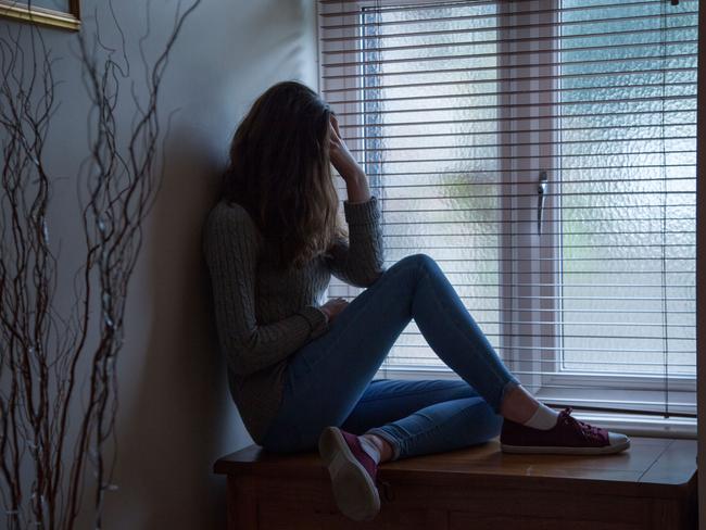 DTR914 Young female with long hair wearing jeans, hand on head in despair, sitting in darkness by a window at home. Side viewpoint.