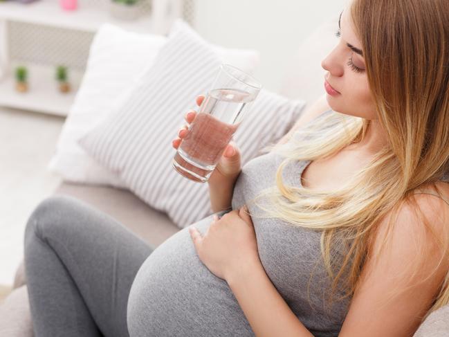 Pregnant woman with glass of water sitting on sofa closeup. Young expectant blonde rest at home. Pregnancy, healthcare, thirst concept Picture: Istock