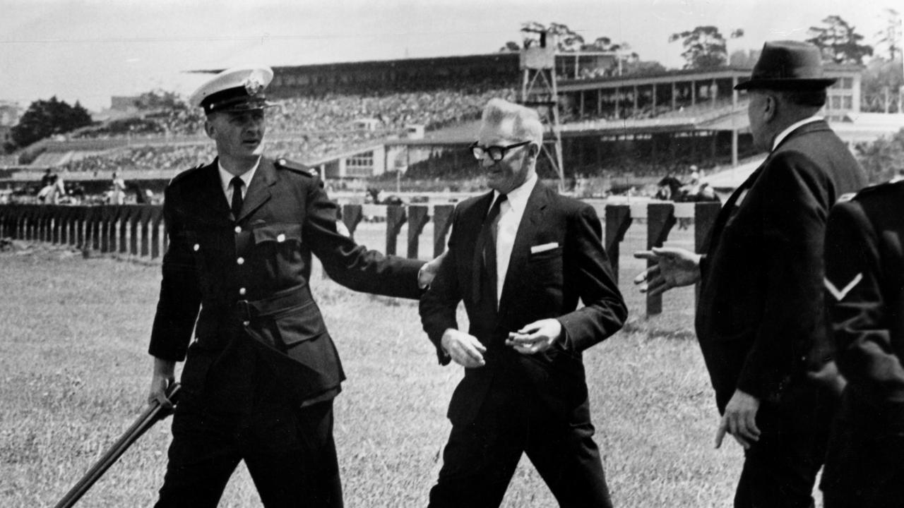 1966. Wally Hoysted is lead away at at Flemington racecourse. Police officer carring the shotgun he had held. Walter Hoysted. Anti-whip crusader. Whipping. Horse racing. Gun. Picture: BRUCE HOWARD Picture: Photo File