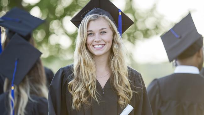 A multi-ethnic group of university students are outdoors at a graduation ceremony. They are wearing graduation robes and caps. They are walking away from the camera, and a Caucasian woman is turned to smile at the camera while holding her diploma.